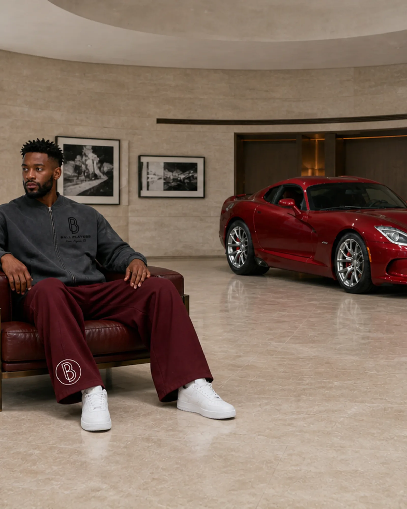 A man models a washed Ball Players jacket and wine-red pants beside a red sports car in a limestone members' garage.