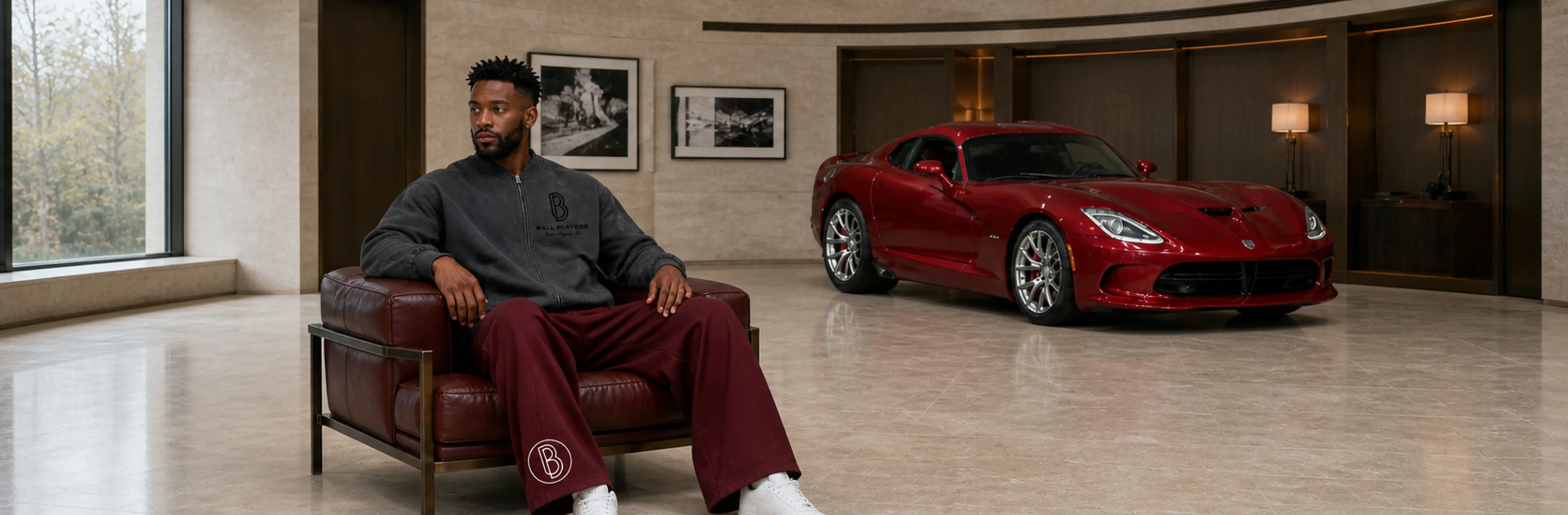 A man wears a washed Ball Players jacket and wine-red sweatpants in a limestone motor salon with a stationary red sports car.